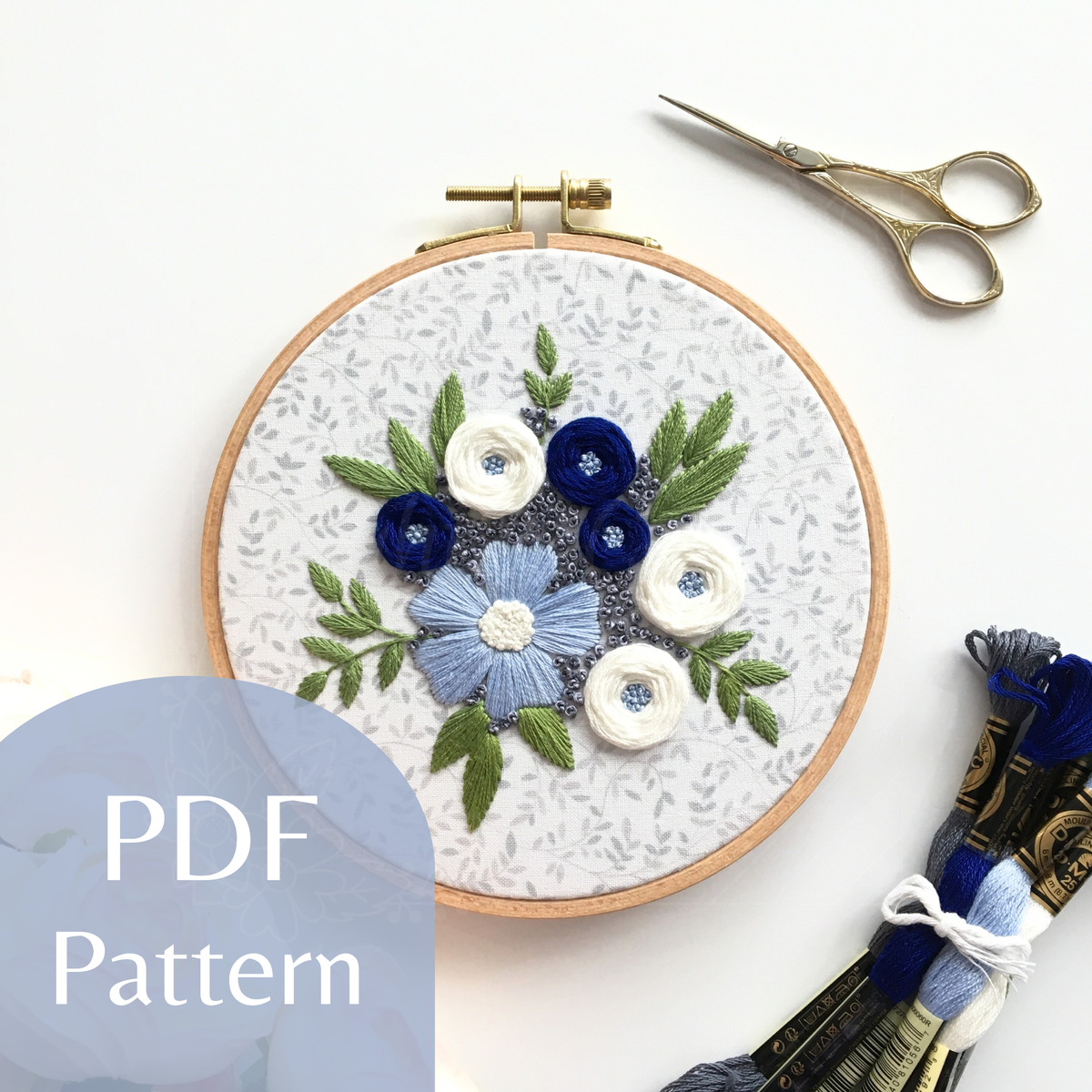 Stitched embroidery design "Winter Blooms" by Eight22Crafts displayed in a round hoop alongside scissors and embroidery thread on a white background