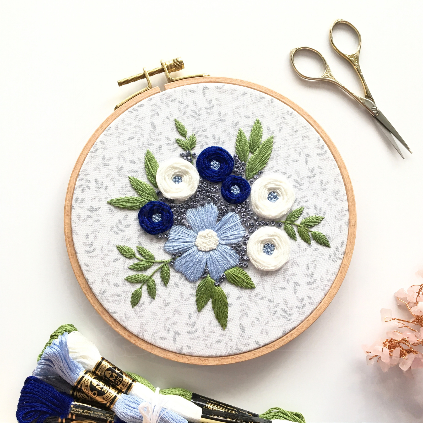 Stitched embroidery design "Winter Blooms" by Eight22Crafts displayed in a round hoop alongside scissors and embroidery thread on a white background
